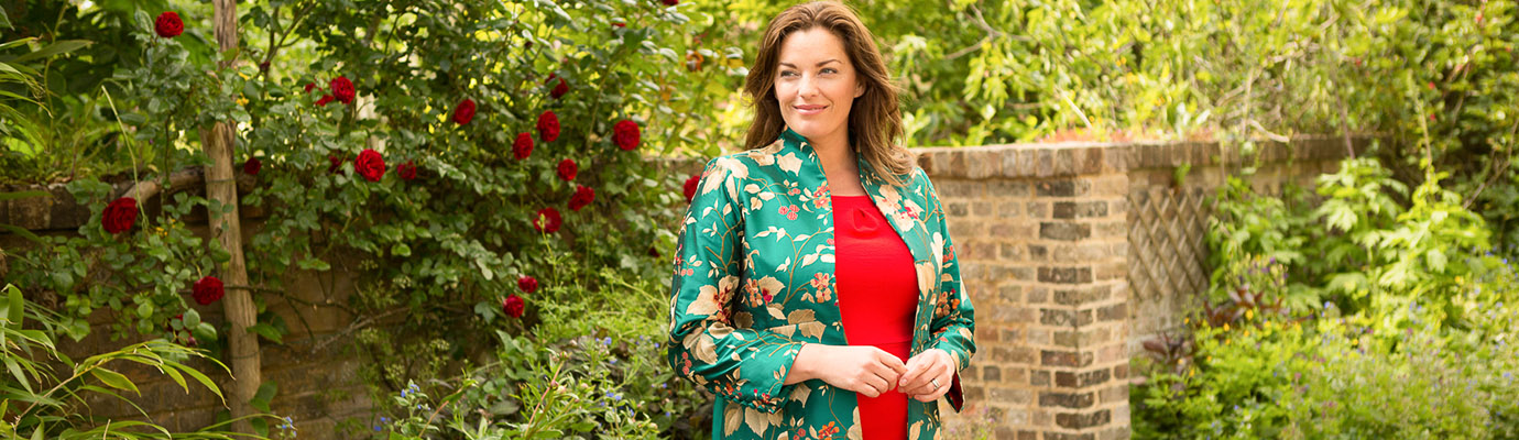 Dark haired woman stands in a garden in a red dress and green embroidered silk coat, the Avani coat by Shibumi.