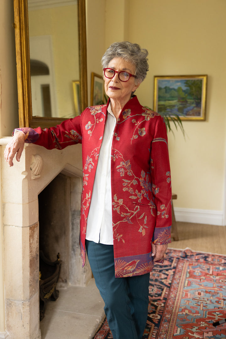 A mature lady standing by a fireplace wearing a striking red jacket with beautiful flower pattern. Elegant outfit for over 70
