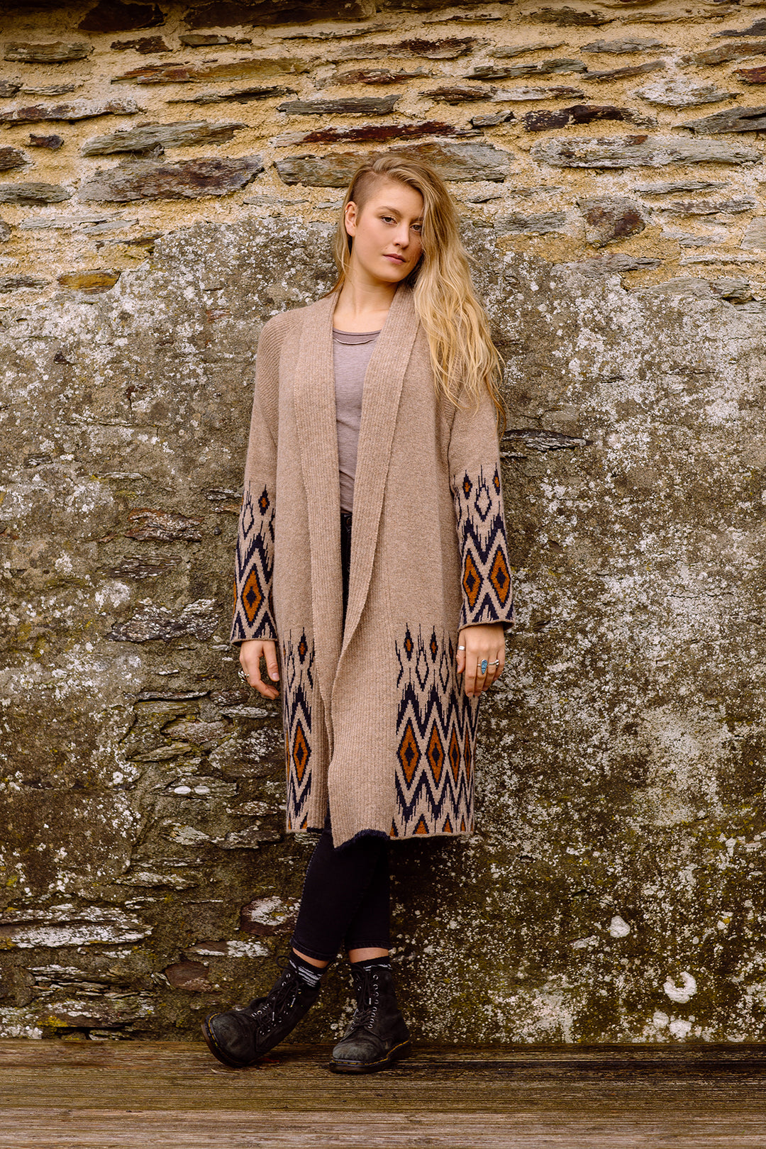 A blond woman with long hair, standing next to a stone wall wearing long wool cardigan.