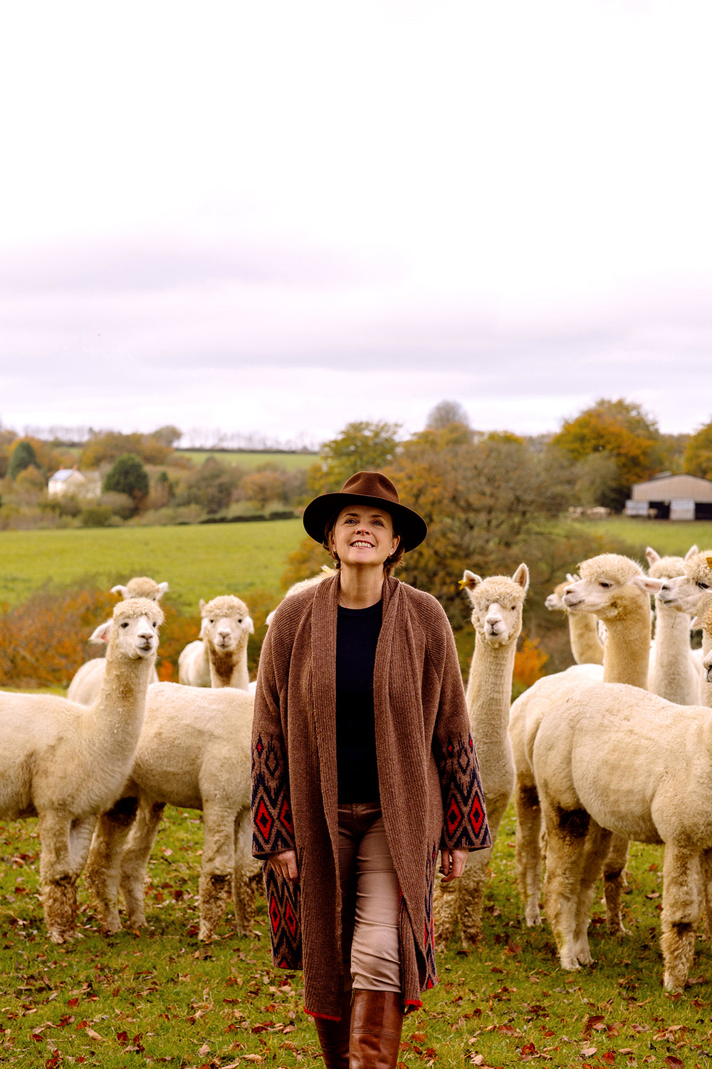 Lady wearing a hat in from of alpacas, with red brown mid length cardigan.