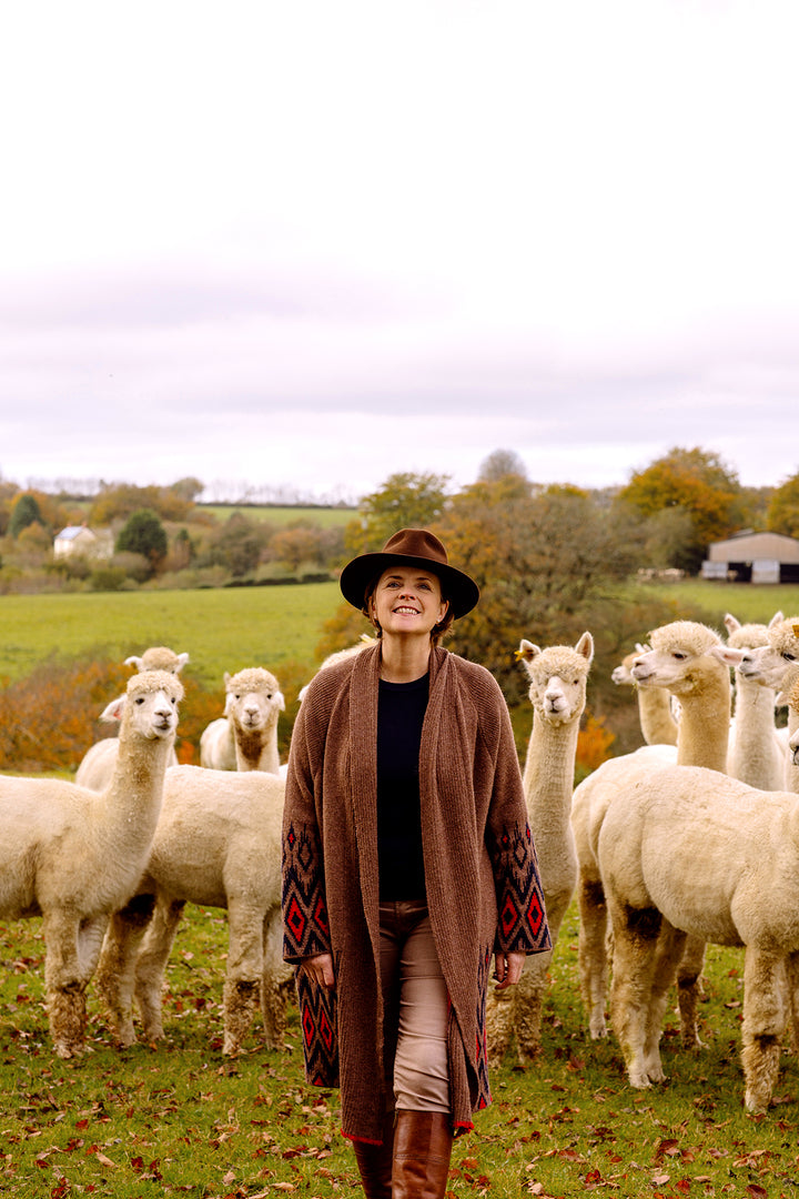 Lady wearing a hat in from of alpacas, with red brown mid length cardigan.
