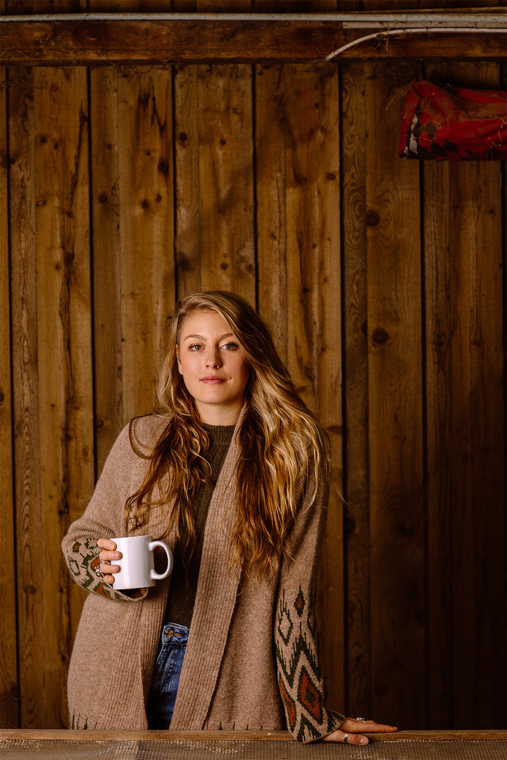Woman with long hair with a cup of tee in her hand, wearing brown cosy cardigan.