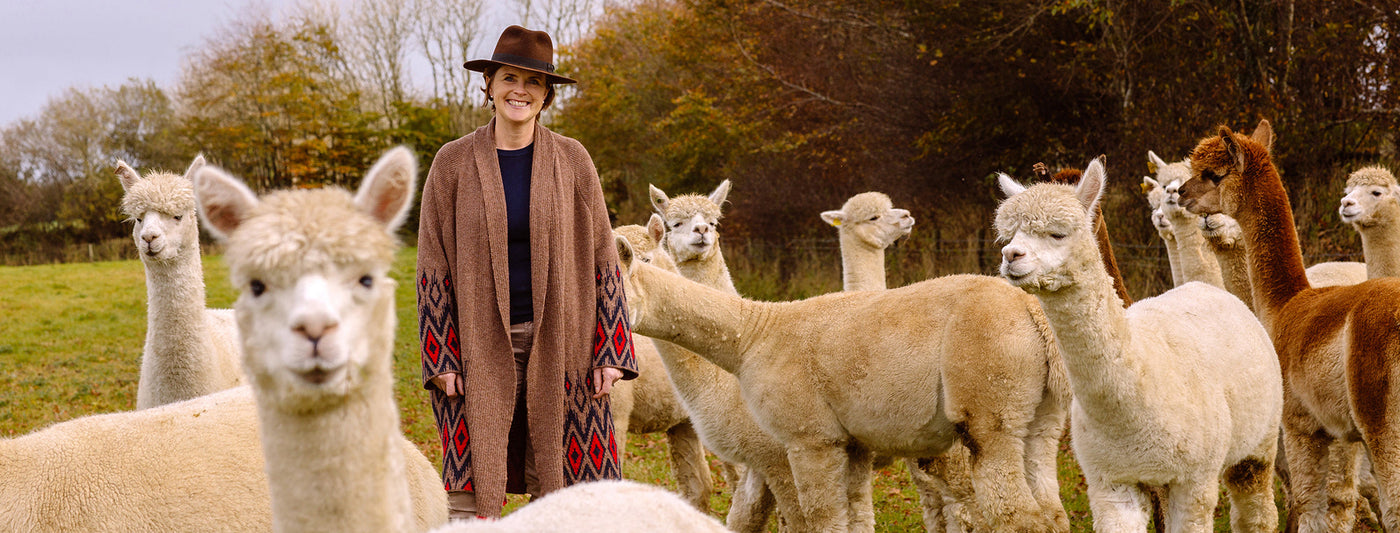 Woman stands in field of alpaca, she wears a cosy women's alpaca cardigan