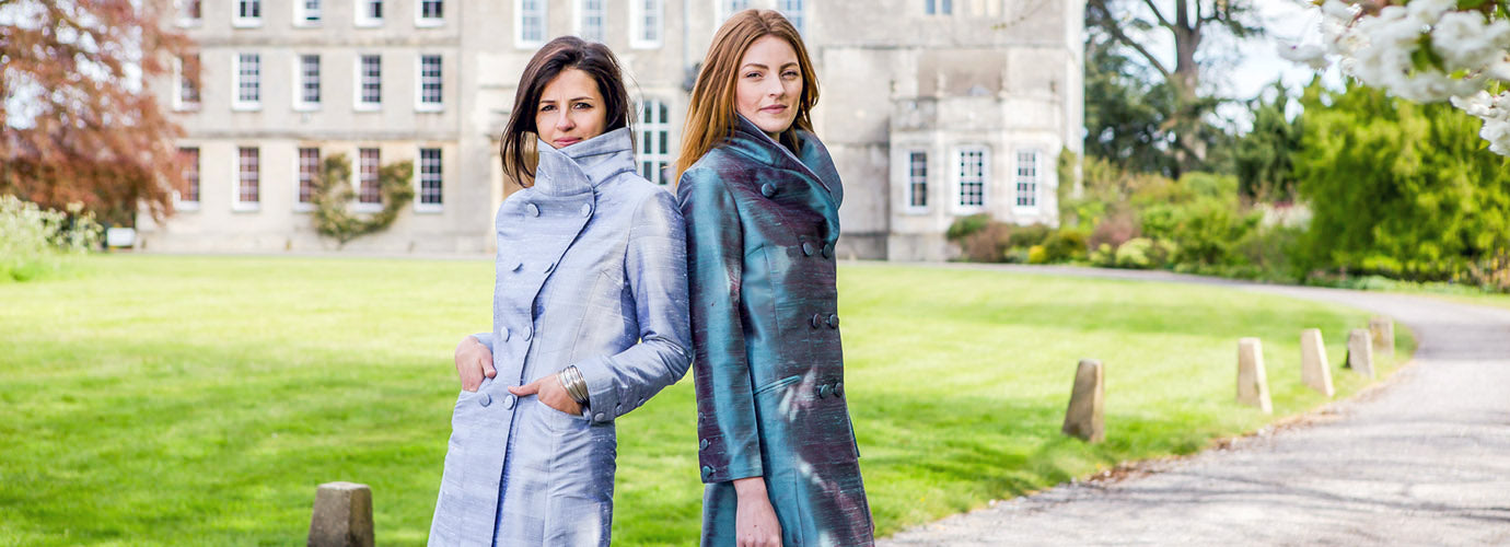 Two women, one red head one brunette in front of stately home both wearing silk coats, the delphine coat in silver and smokey blue