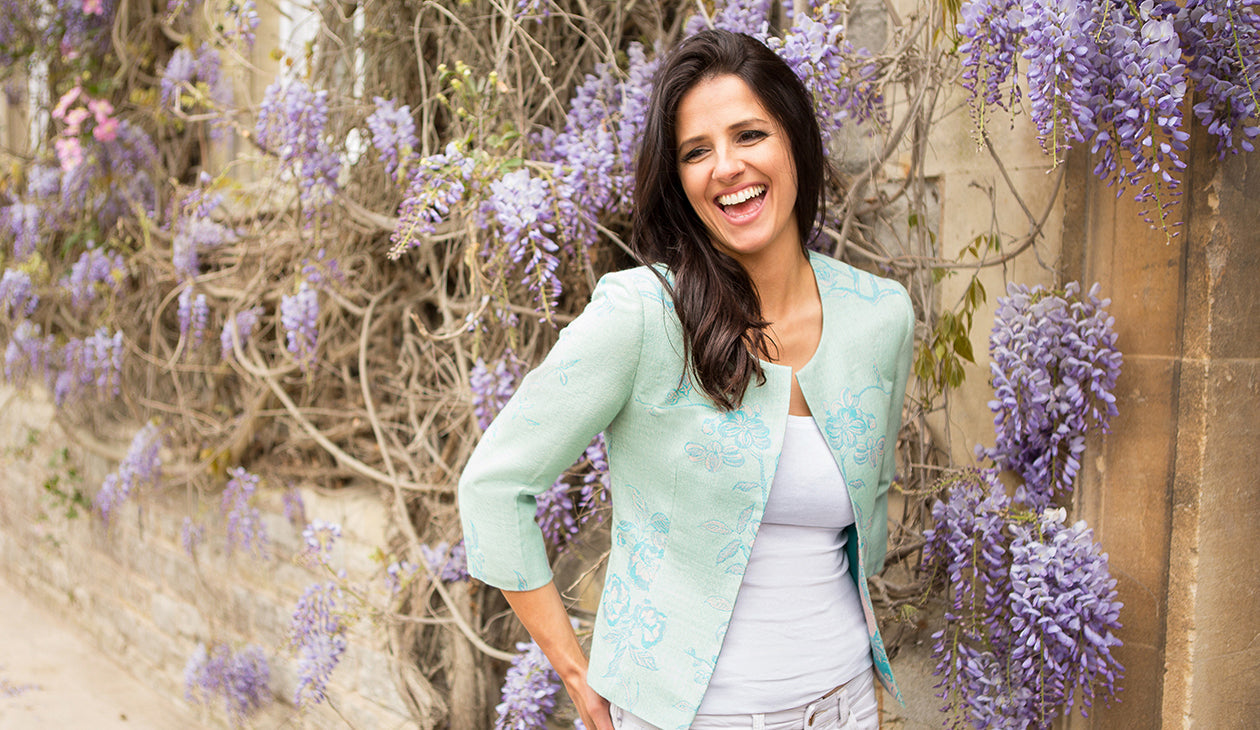 A woman with dark hair laughs in front of a wall of wisteria flowers, she wears a white t-shirt with a pale blue patterned cashmere jacket by Shibumi. 