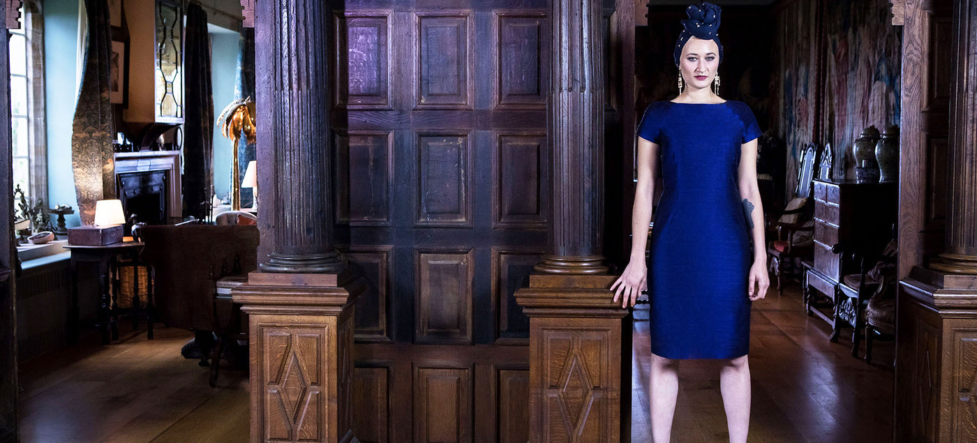 Woman stands in grand panelled room with headscarf wearing a deep navy blue silk shift dress, the Hepburn dress from Shibumi. 