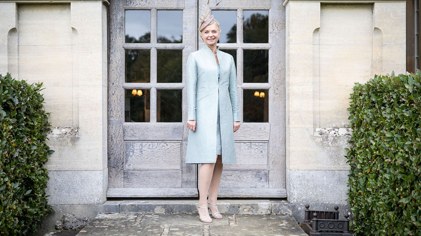A mature woman, mother of the bride or groom or wedding guest wears a pale blue knee length silk Lyra coat with matching dress. 