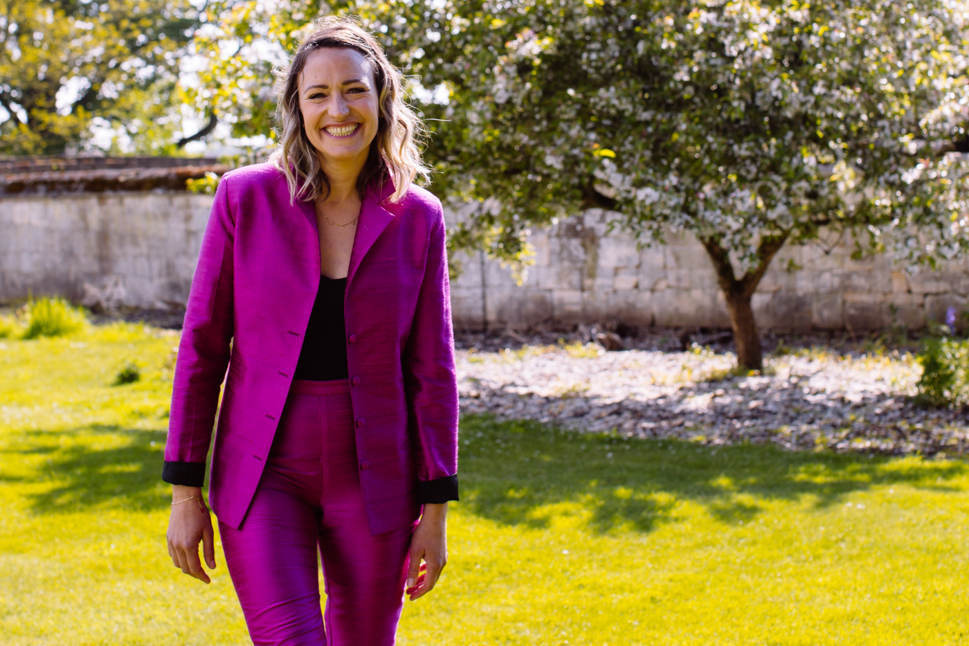 A woman in a bright pink silk suit from Shibumi, cigarette trousers in pink silk paired with a classically tailored silk jacket, ideal women's suit for a wedding guest.