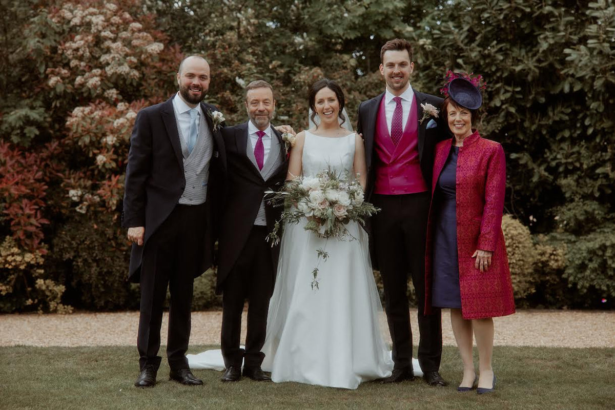A group of five people posing outdoors in formal wedding attire. The bride in a white sleeveless gown holds a bouquet, flanked by two groomsmen in dark suits and a man in a bright magenta vest and tie, with a woman in a textured red shibumi coat and navy dress.