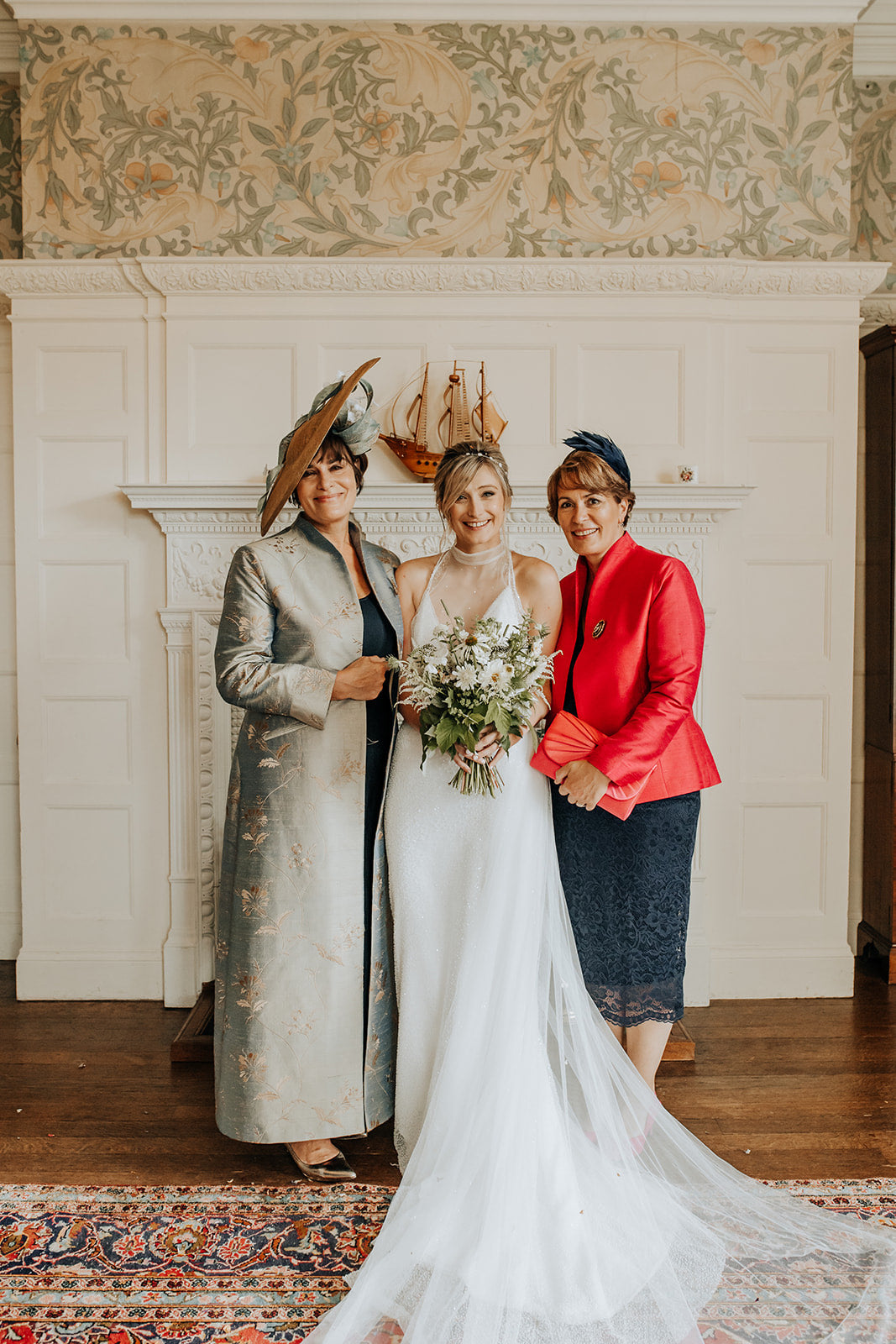 Three women posing together indoors in front of a white fireplace. The central woman wears a white bridal gown and holds a bouquet, flanked by two women in formal attire, one shibumi anya jacket with a hat and the other in a bright red jacket and dark skirt.