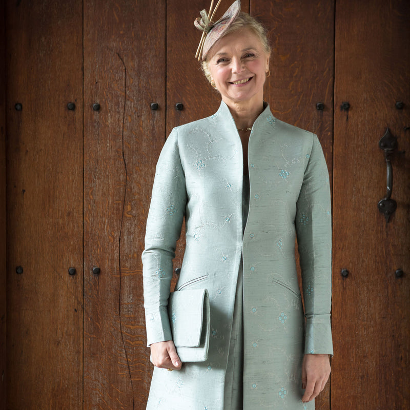 A woman stands in front of a dark wooden door wearing a shibumi lyra coat in moon with a stand collar, matching hat, and fascinator.
