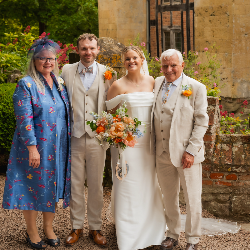 Four people posing outdoors in front of a stone building with brickwork and greenery. The bride in a white off-the-shoulder gown holds a bouquet of orange and peach flowers, flanked by two men in light beige suits and a woman in a blue floral dress and hat.