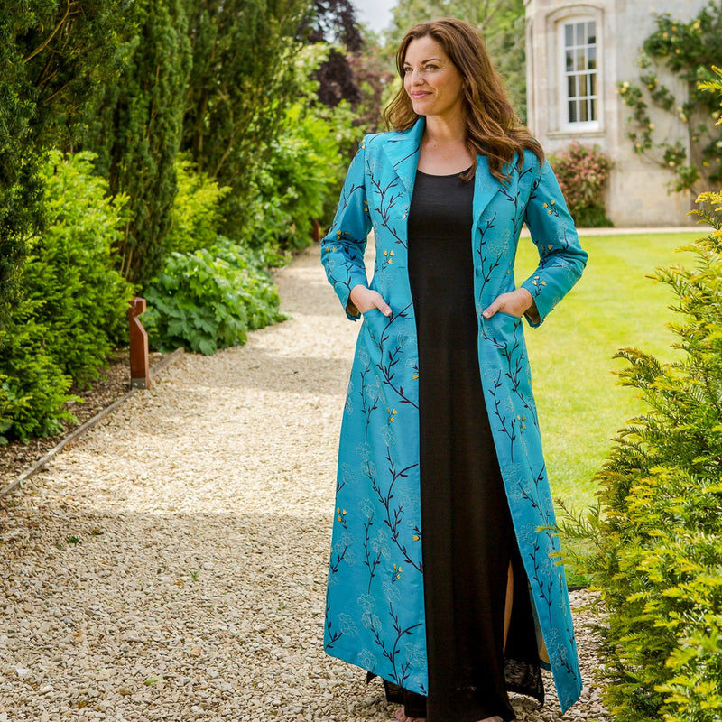 A woman stands on a gravel path in a garden wearing a long turquoise blue floral print coat over a black dress. Trees, shrubs, and a stone building are visible in the background.