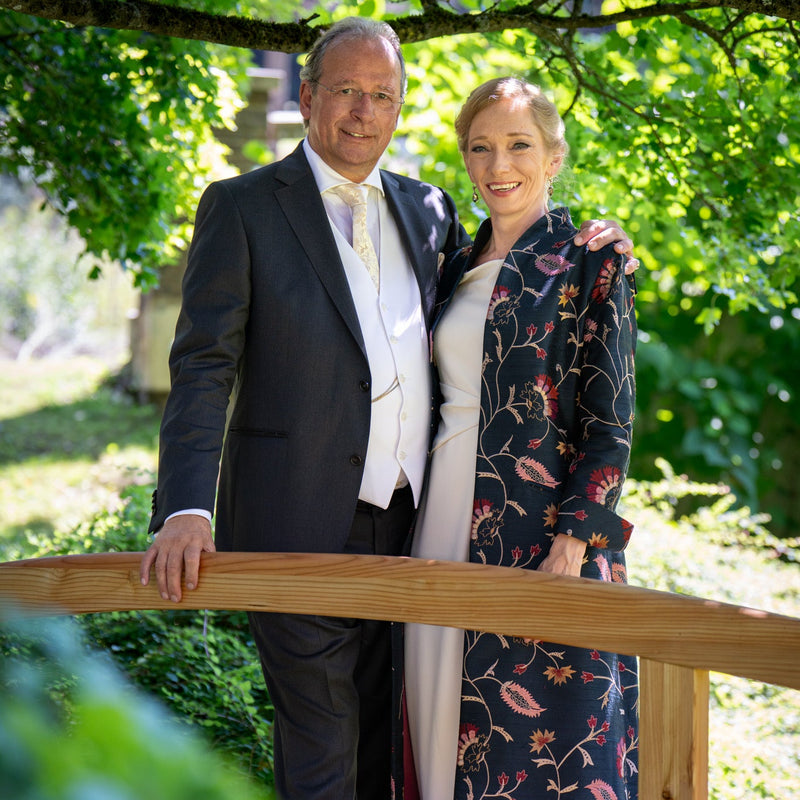 A man in a dark suit and a woman in a floral navy dress standing behind a wooden railing in a garden setting with lush green foliage.