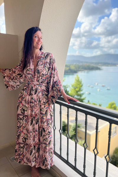 Woman on holiday on a balcony wearing a luxurious pale pink silk velvet dressing gown designed with a beautiful striking botanical pattern embodying many different colours