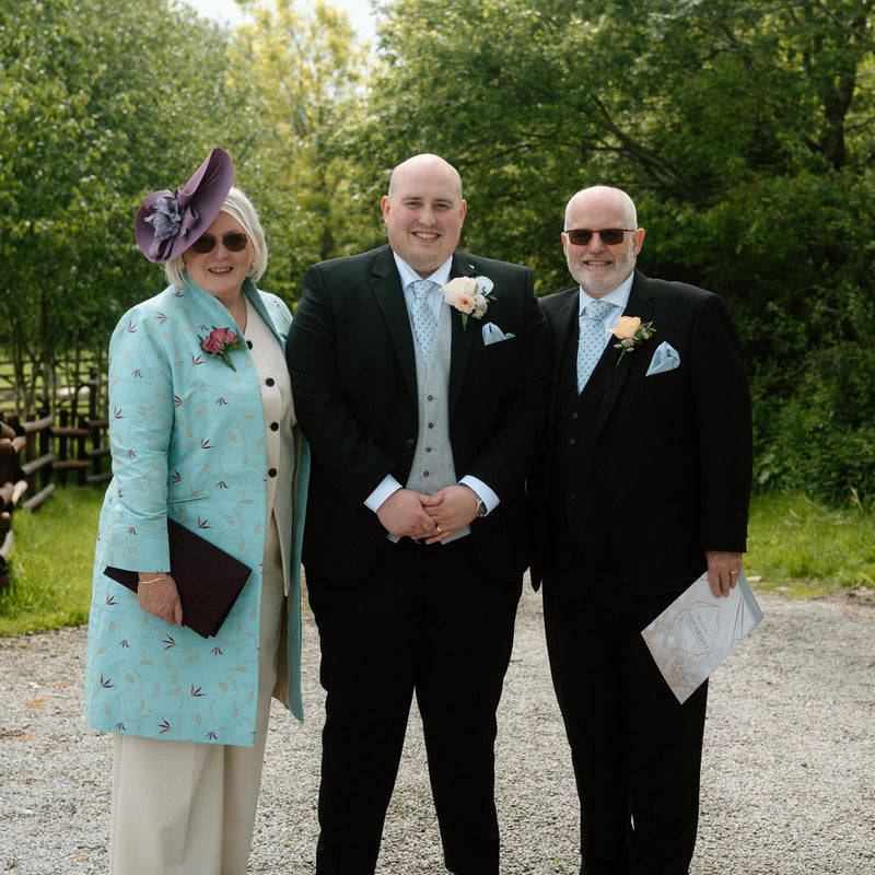 Three people standing together outdoors on a gravel path with trees in the background. The woman on the left wears a light blue patterned coat, cream trousers, and a purple hat, while the two men wear black suits with white shirts and boutonnieres.