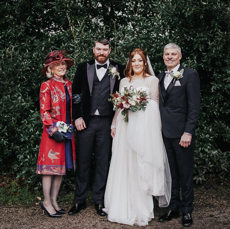 A family portrait of four people standing outdoors against a dense green hedge. The bride in a white wedding gown holds a bouquet, flanked by a groom in a dark suit, an older man in a dark suit, and a woman in a red patterned dress and hat.