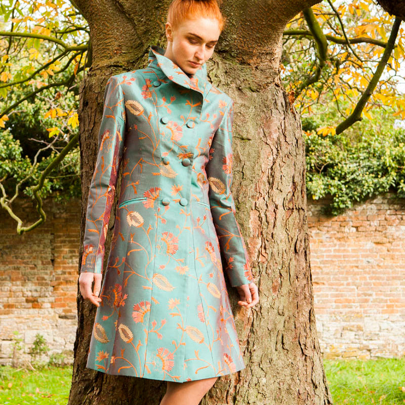 A woman with red hair in a bun stands outdoors leaning against a large tree trunk, wearing a knee-length teal coat with an orange and brown floral pattern and beige heels.