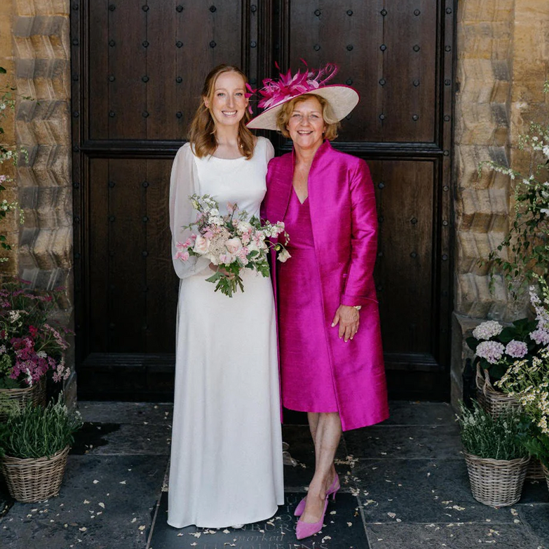 A bride in a white wedding gown holding a pink and white bouquet stands next to her mother, who is wearing a vibrant magenta satin coat, matching pink heels, and a matching pink fascinator hat, posed in front of a dark wooden church door.