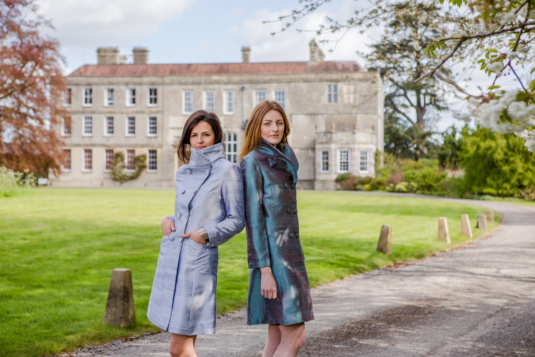 Two women standing on a path in front of a large stone mansion, wearing long high-neck coats. One coat is a light dusty blue-gray, and the other is a dark teal and black patterned coat.