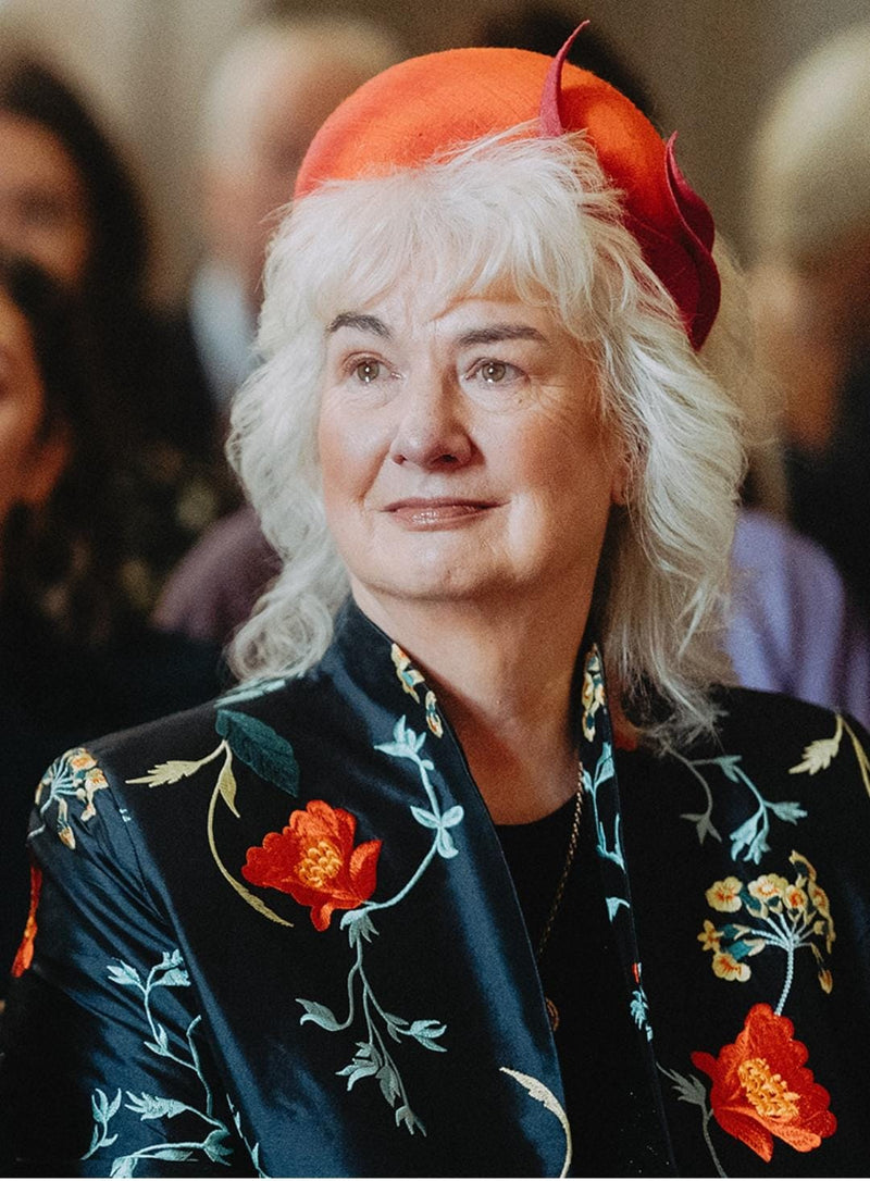 An older woman with white hair wearing a bright orange hat and a dark navy embroidered jacket with red and yellow floral motifs, photographed in a softly blurred indoor setting.