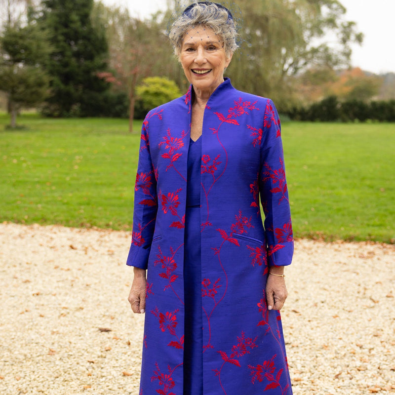An older woman stands outdoors on a gravel path wearing a long, vibrant royal blue coat with a red floral pattern. The background shows a green lawn and trees under an overcast sky.
