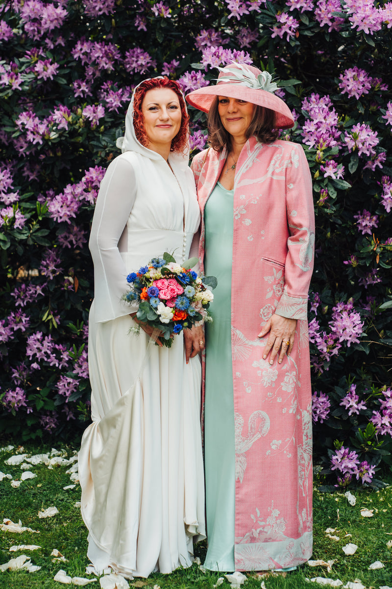 Two women standing outdoors in front of a dense wall of purple flowers. The woman on the left wears a white bridal-style gown with a sheer hood and holds a multicolored bouquet.
