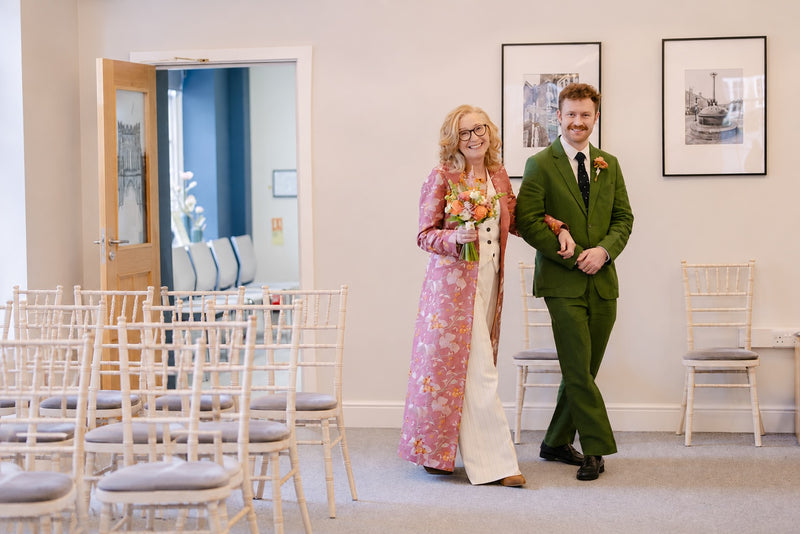 A mother of the groom and groom walk down aisle during their wedding. The bride wears a pink floral longline wedding dress with a cream underlayer and holds a bouquet.