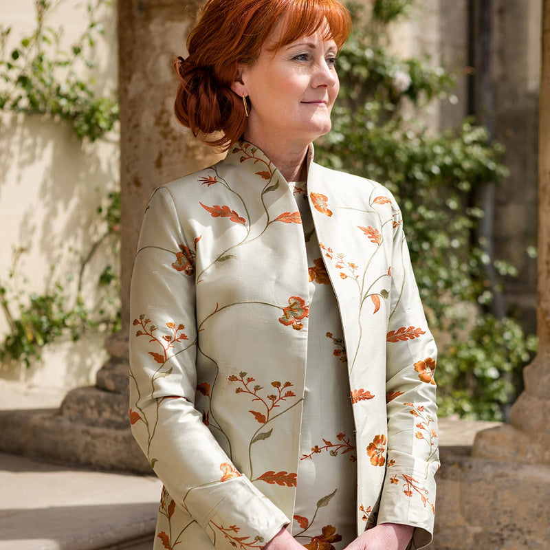 A woman with red hair wearing a cream-colored long coat with an orange and green floral pattern, standing outdoors in front of stone columns and greenery.