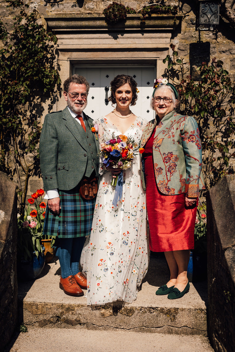 Three people standing together on stone steps in front of a stone building doorway. The central person is a woman in a white floral wedding dress holding a colorful bouquet, flanked by a man in a green blazer and kilt on the left, and an older woman in a red dress and patterned jacket on the right.