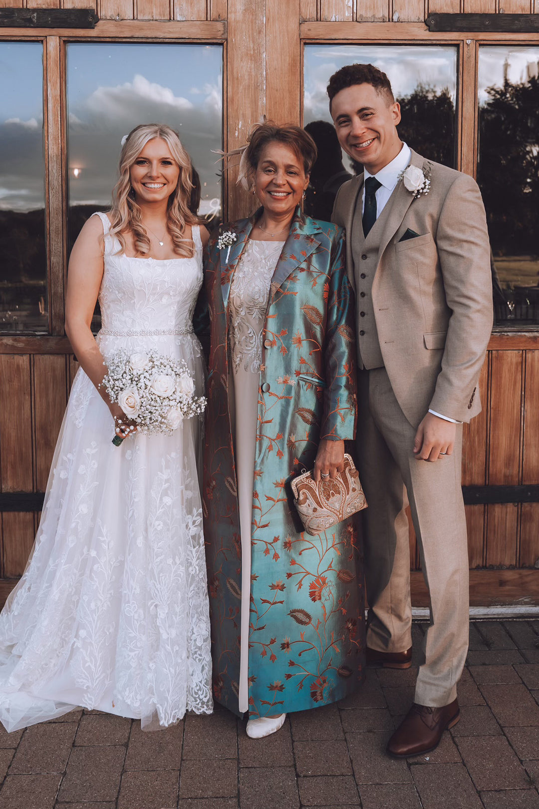 Three people posing together outdoors in front of a wooden building. The woman on the left wears a white lace wedding dress and holds a bouquet, the woman in the center wears a teal floral shibumi  coat over a white dress, and the man on the right wears a light beige suit with a white boutonniere.