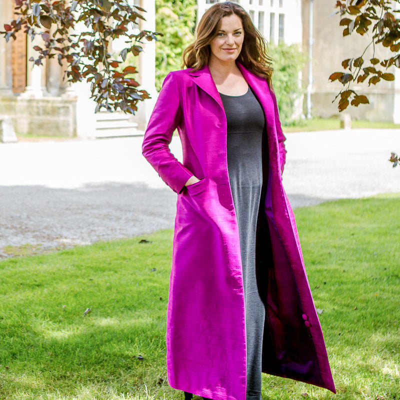 A woman stands outdoors on a grassy lawn in front of a stone building, wearing a long bright magenta coat over a dark gray dress.