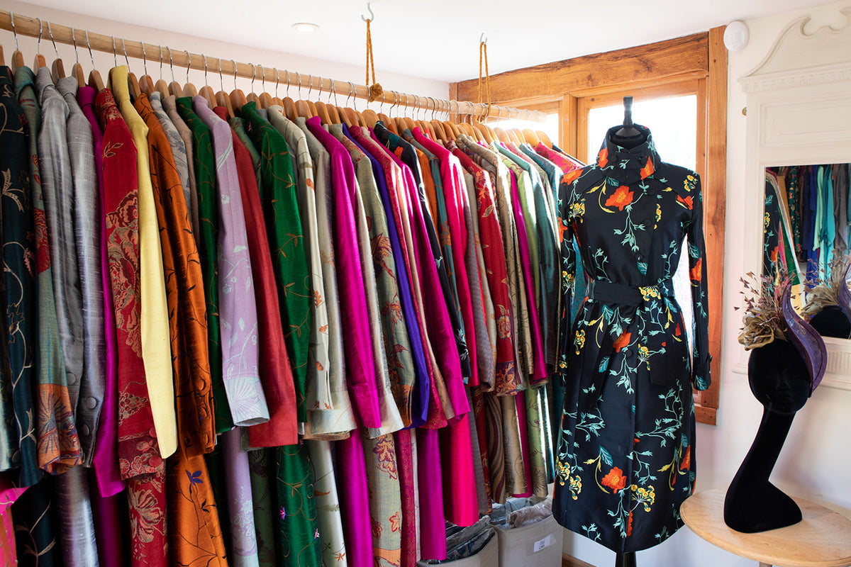 A boutique clothing display at the shibumi showroom featuring a rack of colourful patterned garments on the left and a black long-sleeve floral dress on a mannequin to the right, set in a shop with wooden trim and a mirror.