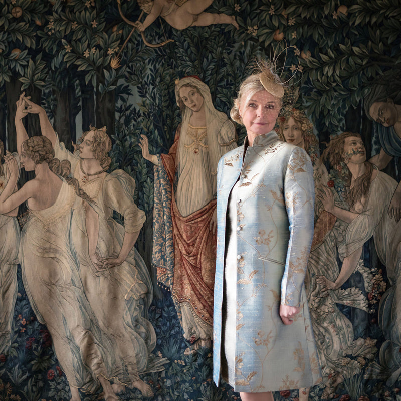 A woman stands in front of a large, detailed tapestry depicting classical figures and a forest scene. She is wearing a light blue, long-sleeved dress with gold floral embroidery and a matching light blue hat with gold detailing.