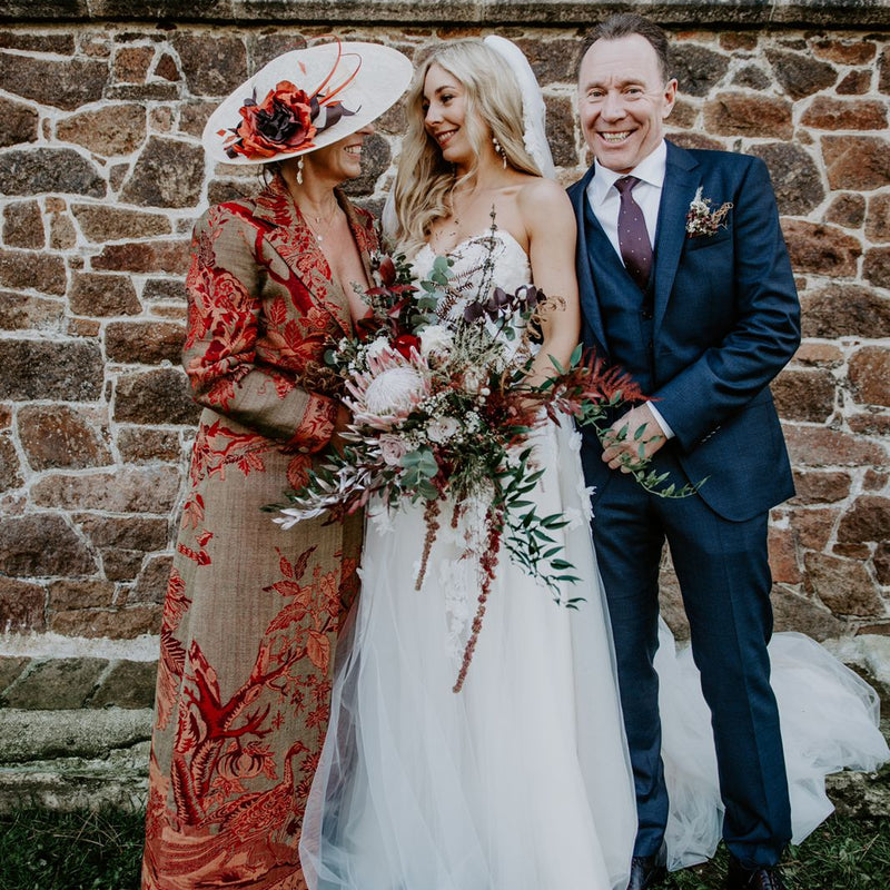 A bride in a white wedding gown holding a floral bouquet stands between her mother, who wears a rust-red embroidered dress and hat, and her father in a navy suit, posing in front of a rustic stone wall.