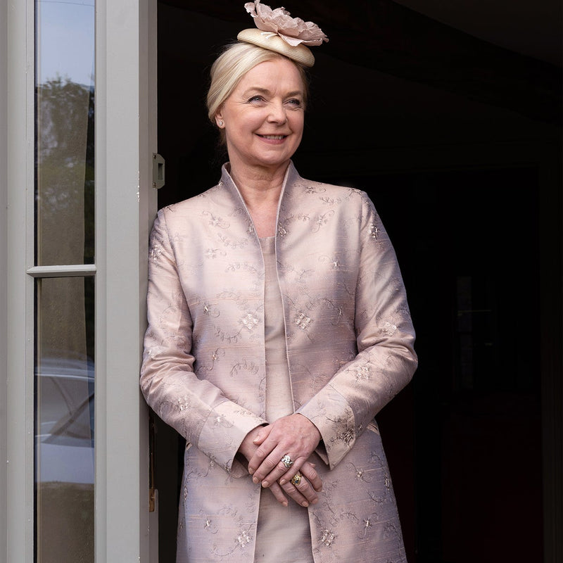 A woman stands in a doorway wearing a light blush pink embroidered long coat over a matching skirt, with a matching pink fascinator hat. The setting appears to be a home entrance with a dark interior behind her.