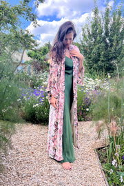 Woman in botanical garden surrounded by blooming flowers wearing a luxurious pale pink silk velvet dressing gown designed with a beautiful striking botanical pattern embodying many different colours
