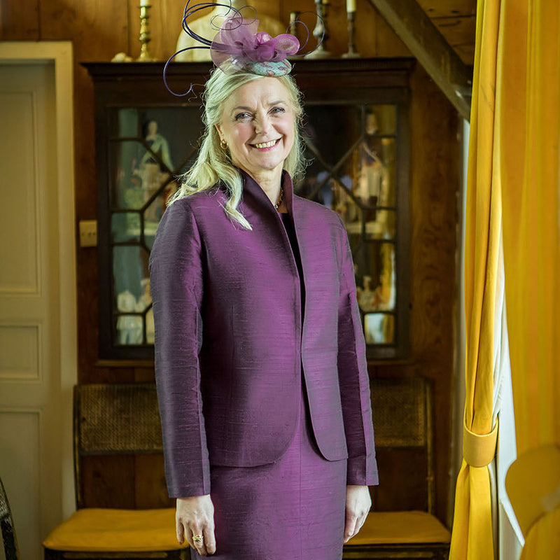 A woman stands indoors wearing a tailored purple skirt suit with a matching fascinator hat. The setting is a warm wood-paneled room with furniture and yellow curtains visible in the background.