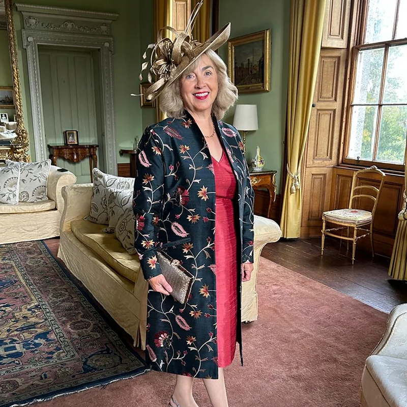 A woman stands in an elegant vintage-style room wearing a dark navy floral coat over a bright pink dress, accessorized with a large decorative hat and holding a metallic clutch.
