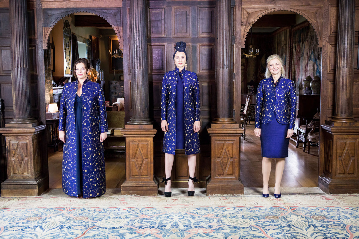 Three women model matching navy blue patterned shibumi outerwear and skirt sets in an ornate wood-paneled room with arched doorways and a patterned rug.