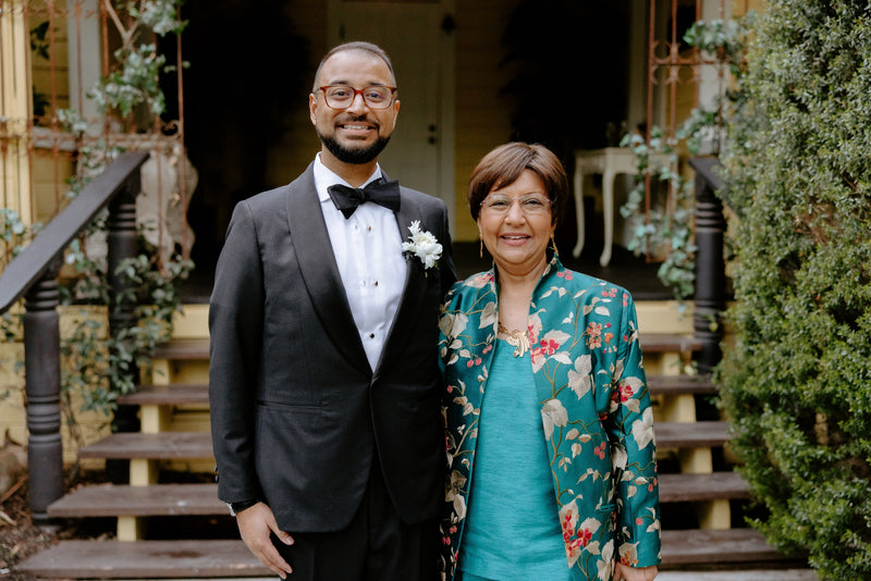 A man in a black tuxedo with a white boutonniere stands next to a woman wearing a teal dress and a green floral sil embroidered shibumi jacket, posing on the wooden steps of a house.