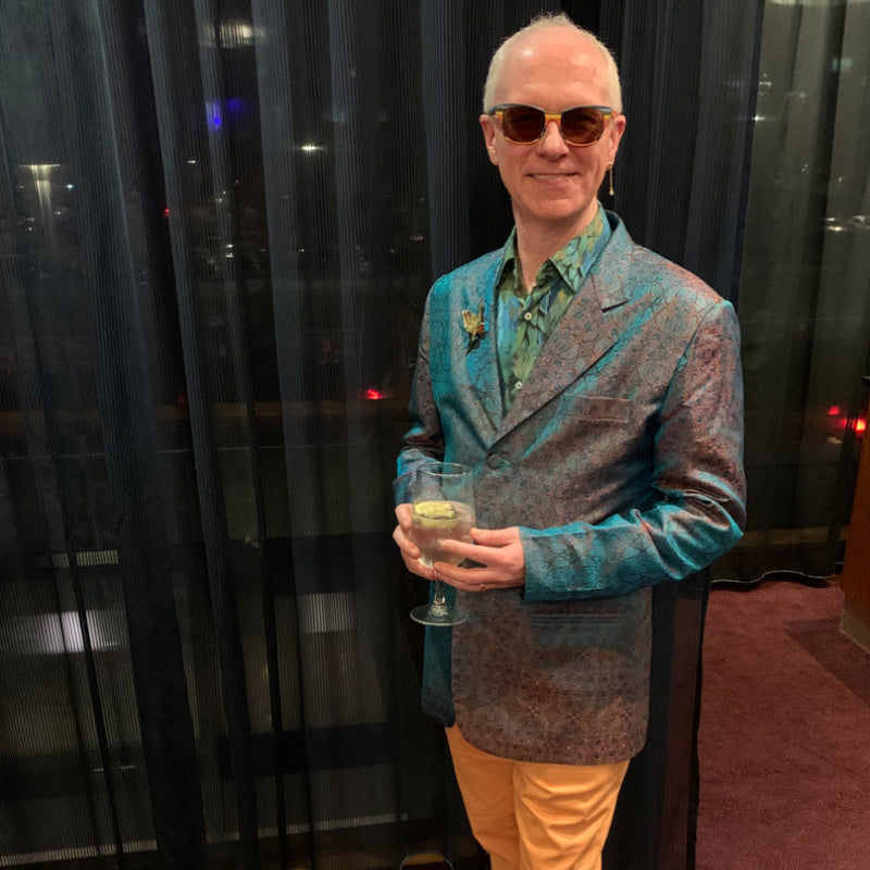 A man wearing a patterned teal and brown blazer, a green shirt, mustard yellow trousers, and sunglasses, holding a glass of champagne in front of dark curtains.