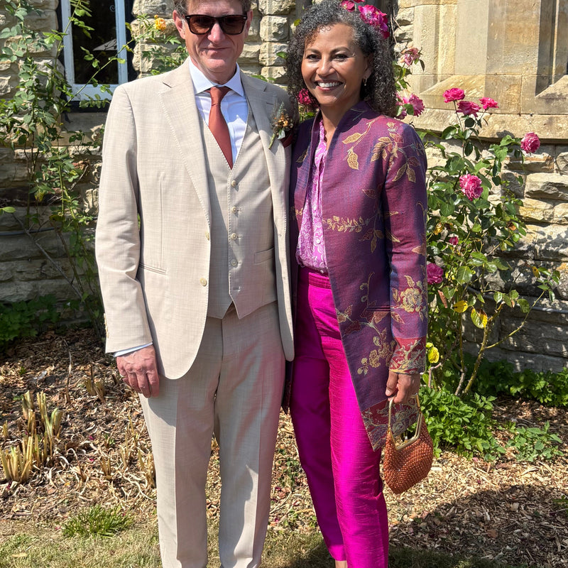 A man and a woman standing outdoors in front of a stone building. The man wears a light beige three-piece suit with a white shirt and a rust-orange tie.