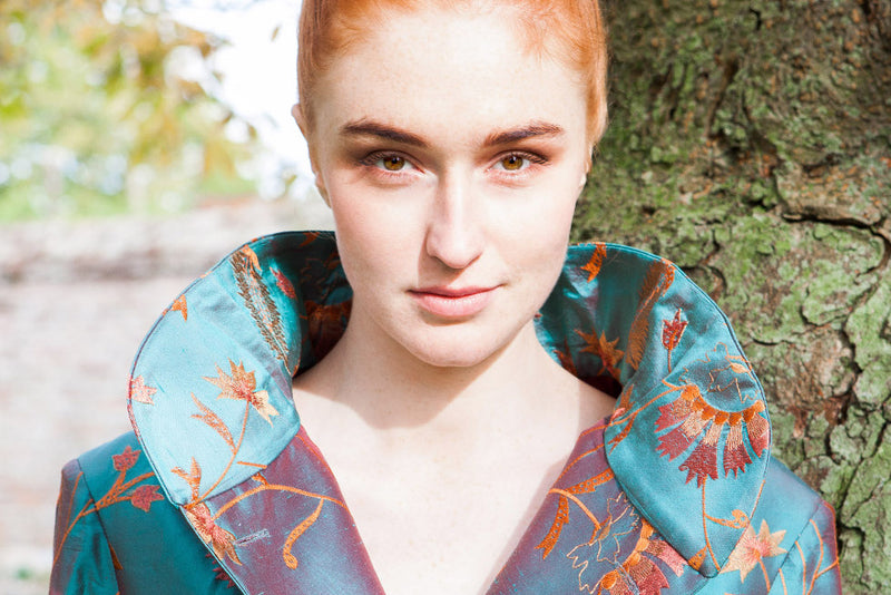 A close-up portrait of a woman with red hair wearing a teal silk shibumi jacket with a large collar and orange floral embroidery, standing outdoors next to a tree trunk.