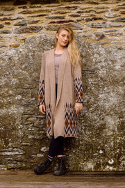 A blond woman with long hair, standing next to a stone wall wearing long wool cardigan. 
