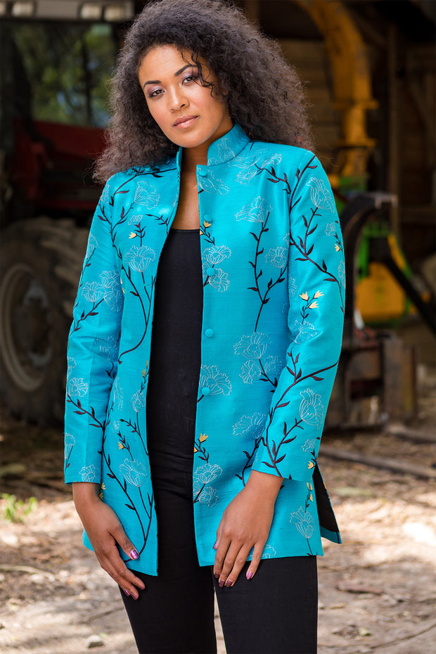 Woman looking straight on wearing a smart, mandarin collar nehru jacket with vents on either side, in a bright turquoise embroidered raw silk with a dark aubergine, gold and very pale grey pattern